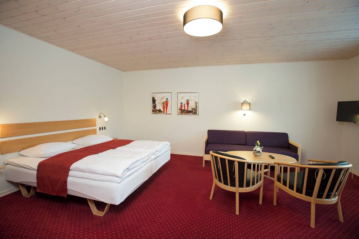 A hotel room with a bed, a sofa, a dining table with chairs, and a flat-screen TV mounted on the wall.
