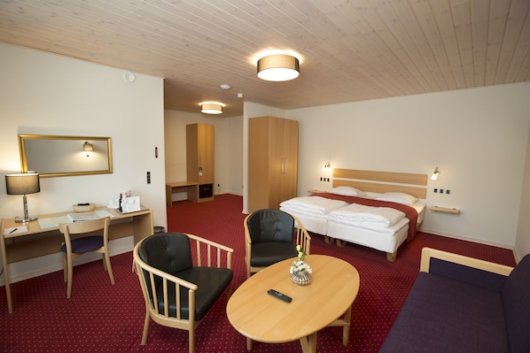 Quarto de hotel com escrivaninha de madeira, duas cadeiras, cama com roupa branca, mesa redonda com um controle remoto e um sofá.