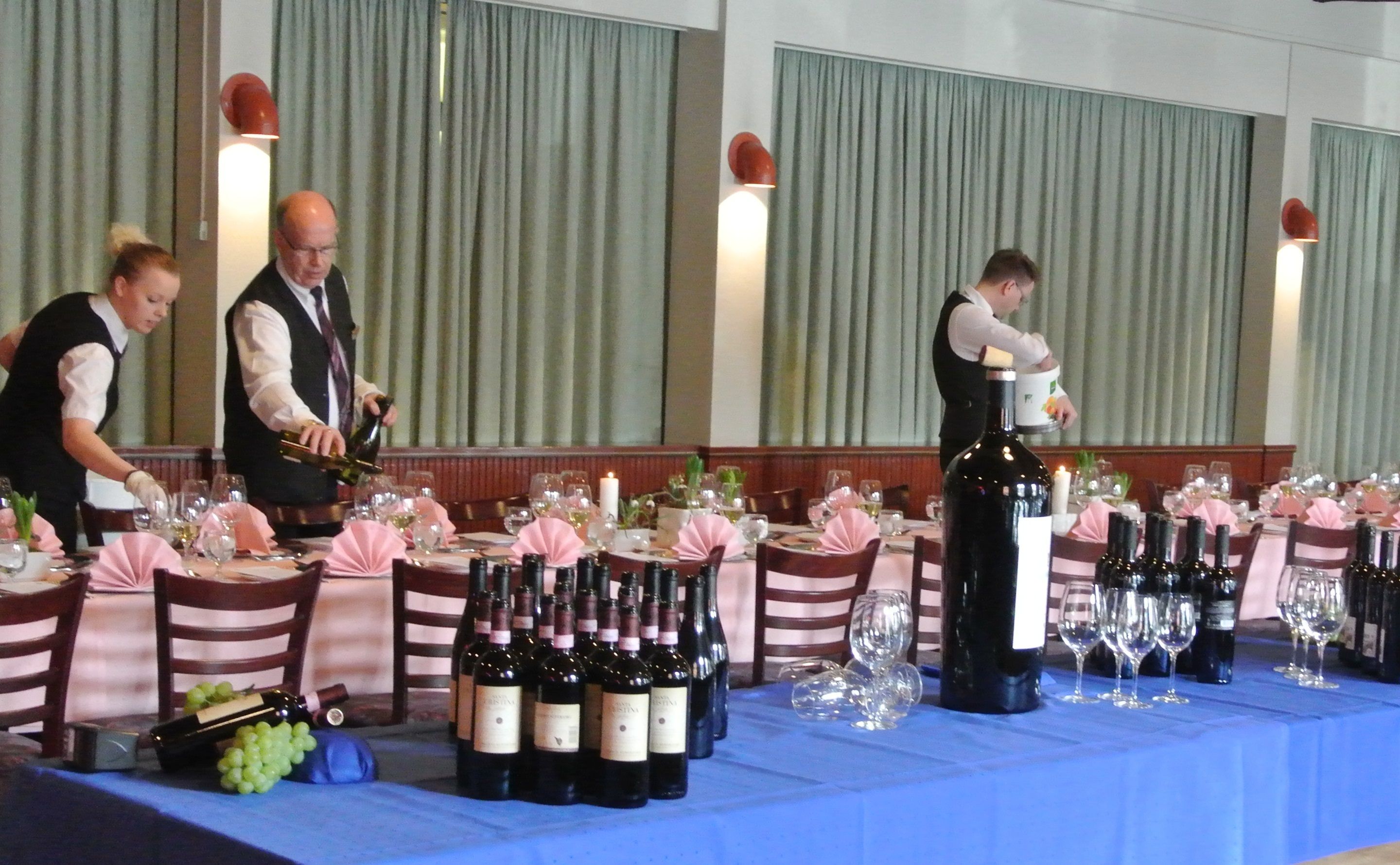 A formal dining setup with multiple tables, each set with wine bottles, glasses, and waitstaff serving.