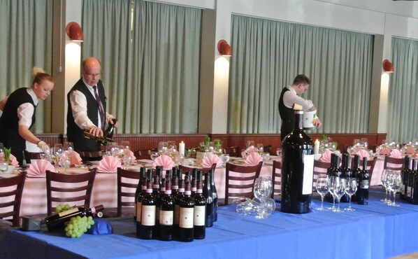 Montaje formal para cena con varias mesas, cada una dispuesta con botellas de vino, copas y personal de servicio.