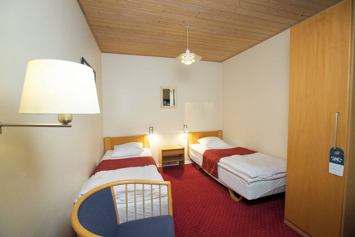 A hotel room with two single beds, a wooden ceiling, a red carpet, a desk, and a chair.