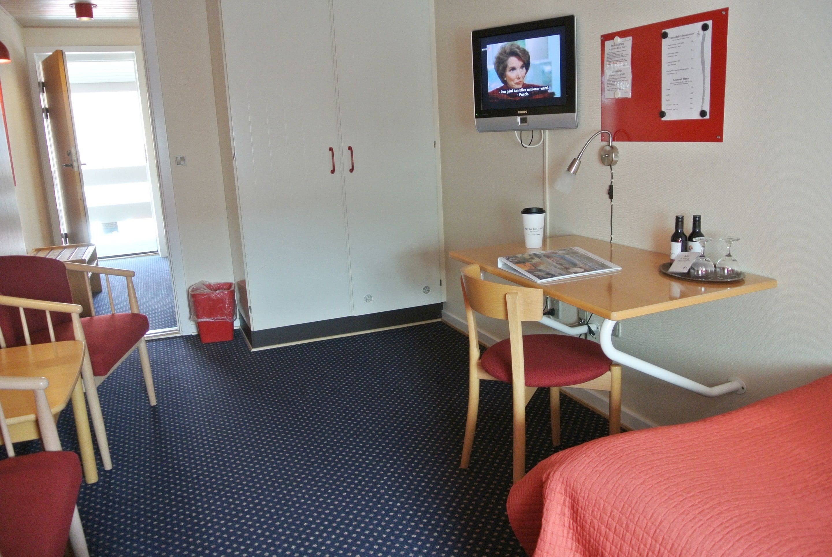 Quarto de hotel com cama, escrivaninha com cadeira, televisão, mural e pequena mesa com copo e garrafas.