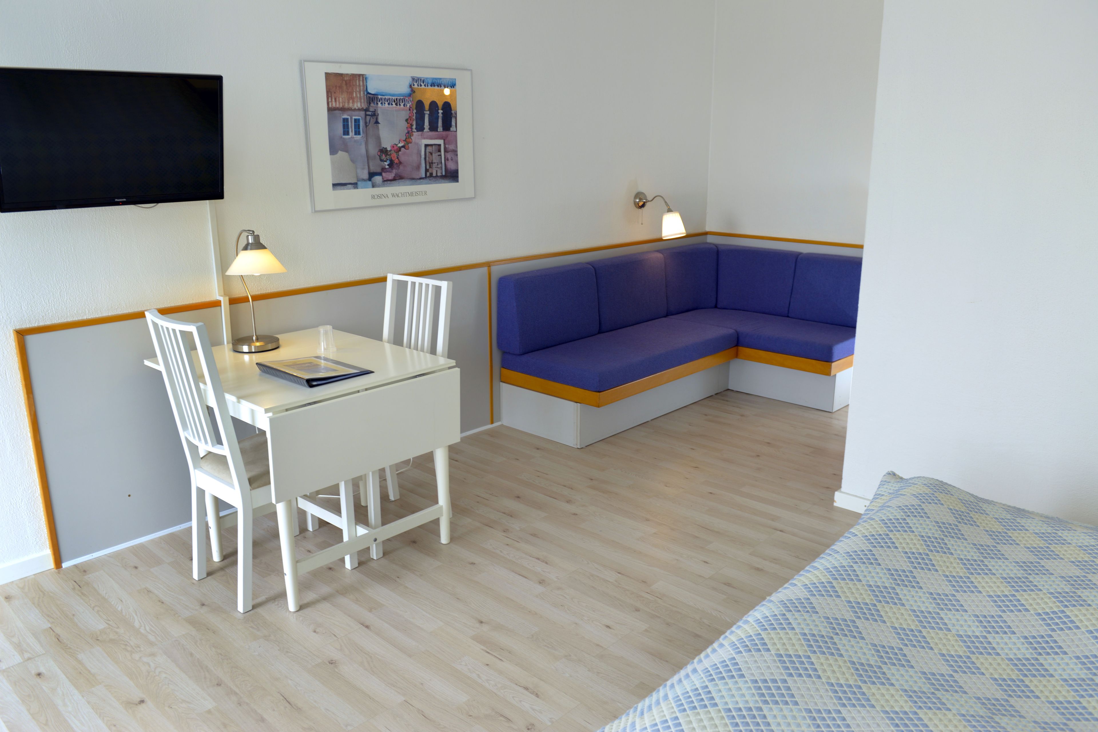 A room with a white table, chairs, a blue sofa, a bed with a patterned cover, a TV mounted on the wall, and a framed picture.