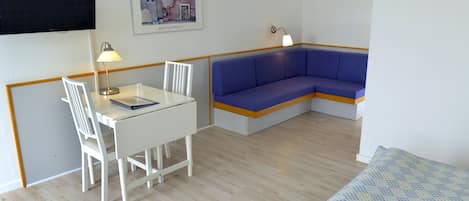 Una habitación con una mesa blanca, sillas, un sofá azul, una cama con cubrecolchón estampado, un televisor montado en la pared y un cuadro enmarcado.