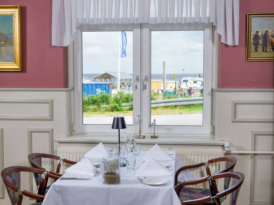 Comedor con una mesa dispuesta para cuatro personas, dos sillas a cada lado, una ventana con vistas al exterior y una cortina blanca sobre la ventana.