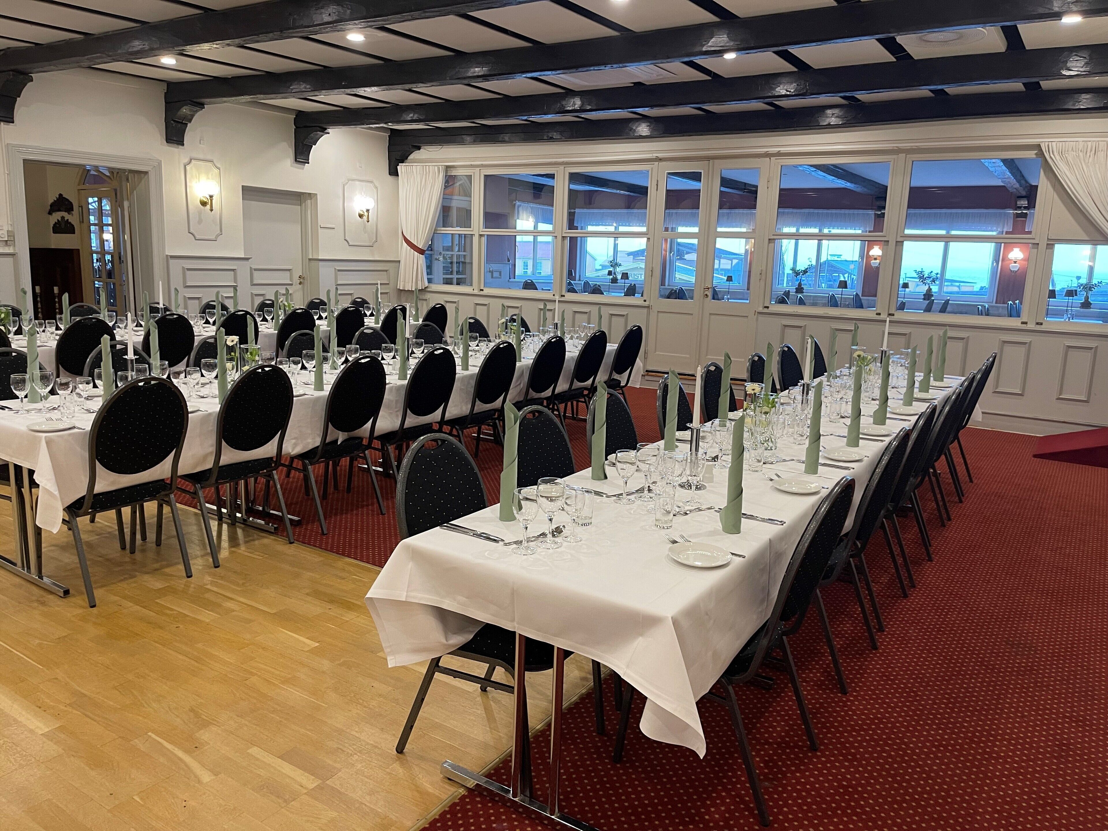 A banquet hall with multiple tables set for dining, featuring white tablecloths, black chairs, and green centerpieces.