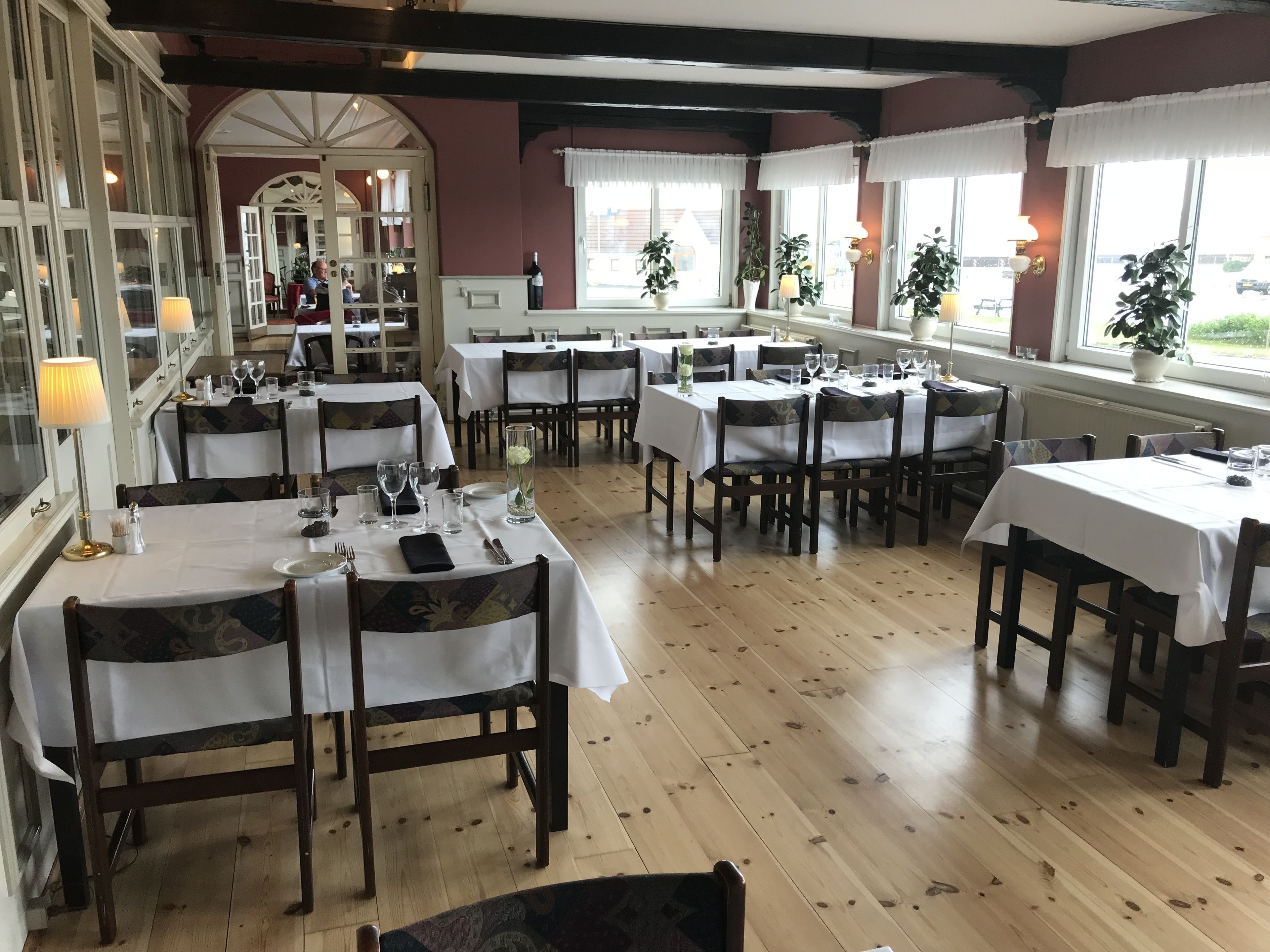 Un restaurante con suelos de madera, manteles blancos y sillas de madera oscura. Las mesas están dispuestas con copas de vino y platos.