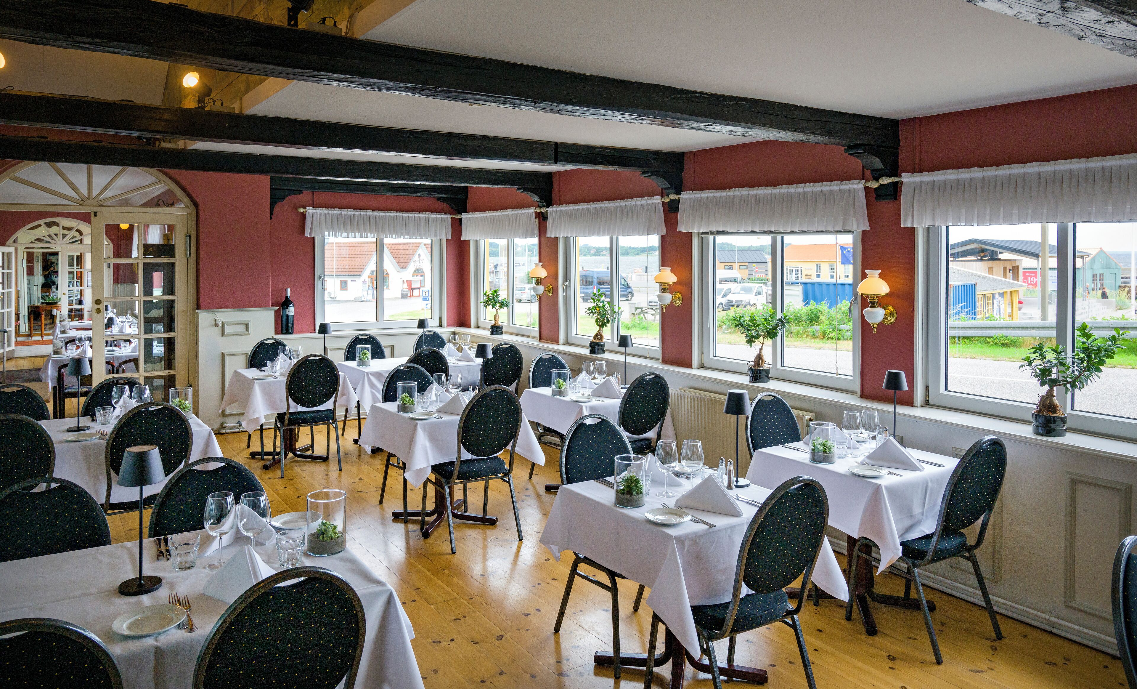 Un restaurante con varias mesas preparadas para comer, amplios ventanales con cortinas y vistas al exterior.