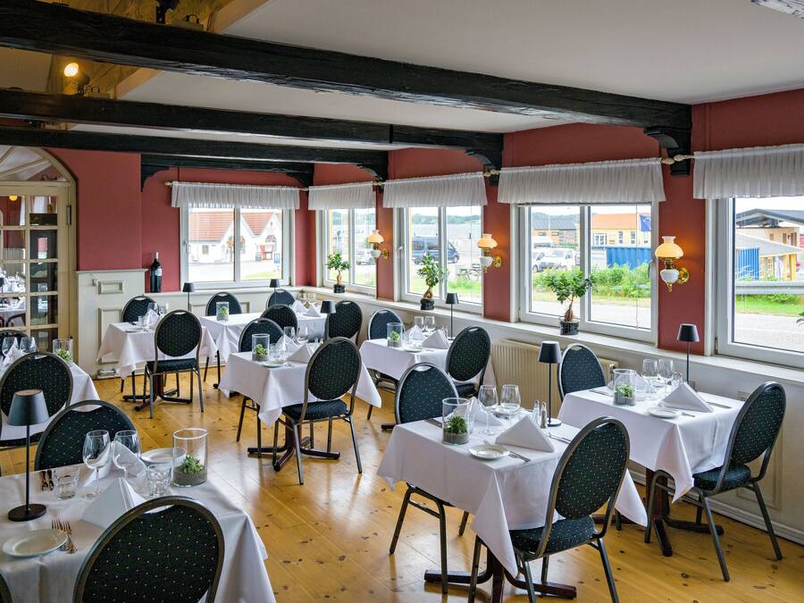 En restaurant med flere spiseborde, store vinduer med gardiner og udsigt ud.