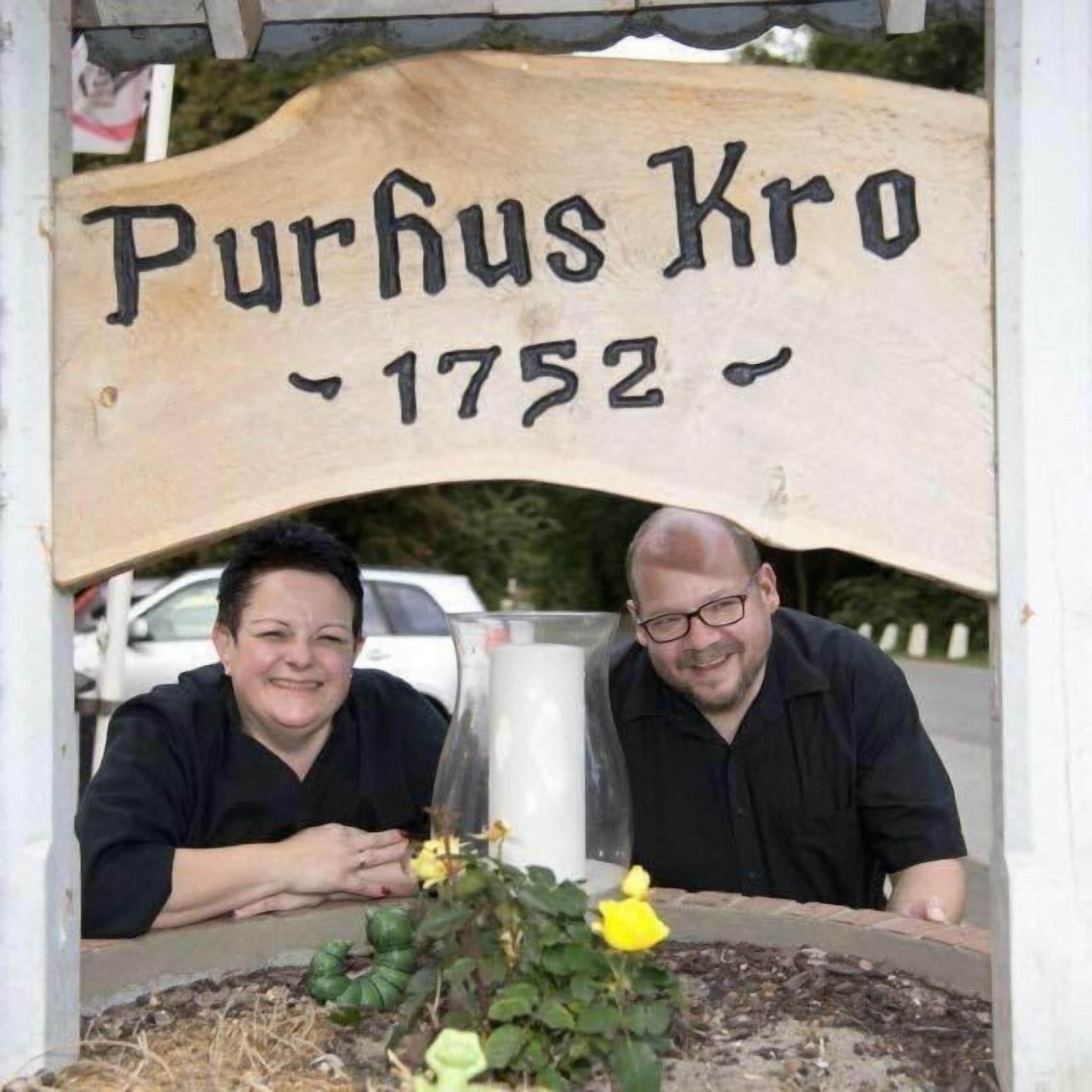 Ein hölzernes Schild mit der Aufschrift 'Purhus Kro 1752' über einem Blumenbeet.