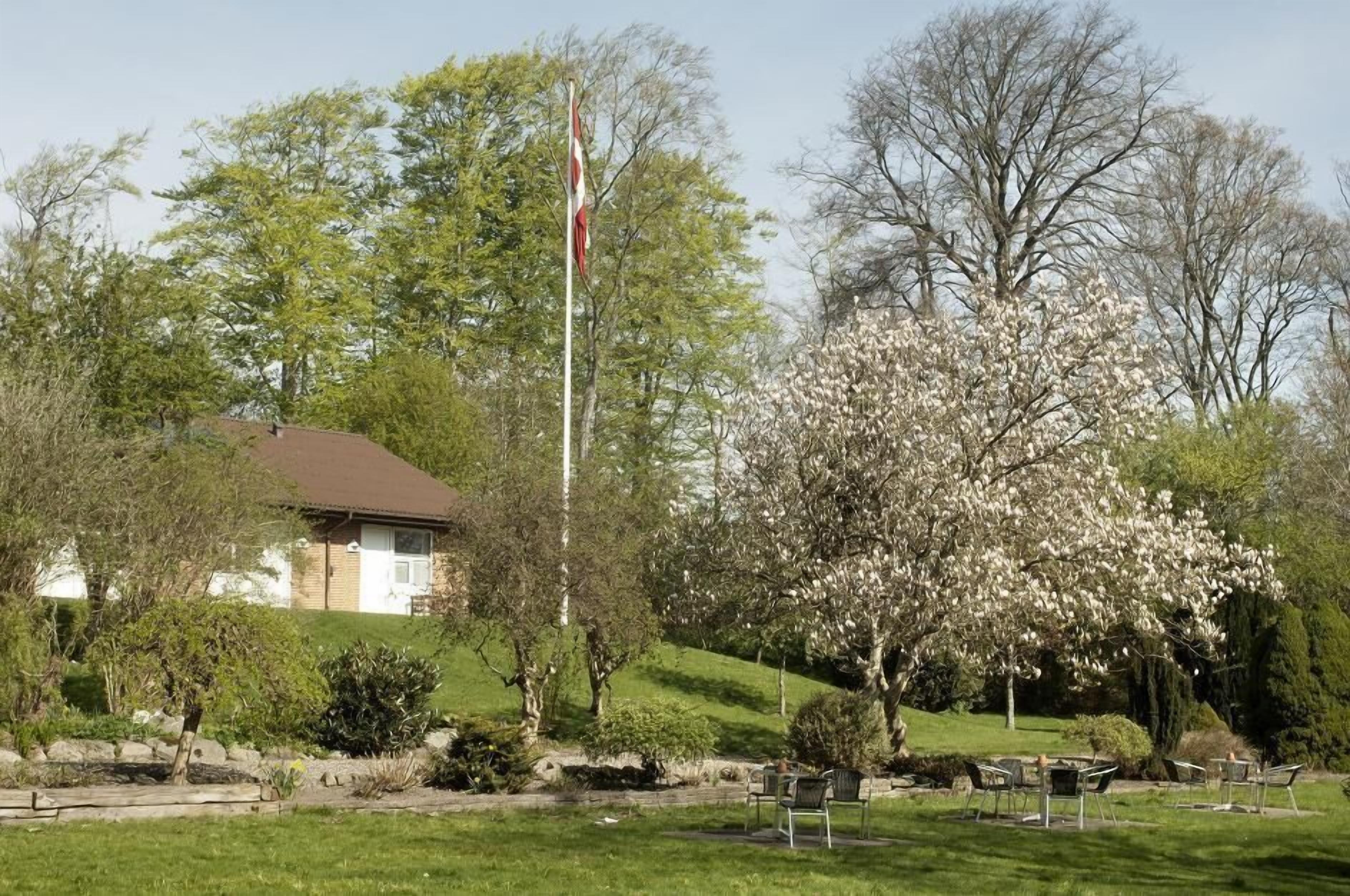 Une maison avec un mât sur lequel flotte un drapeau, entourée d’arbres en fleurs et d’une pelouse aménagée avec des sièges pour l’extérieur.