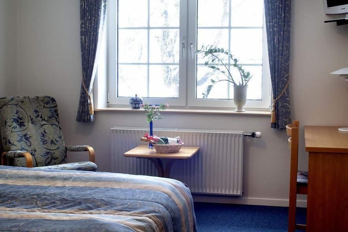 Une chambre d’hôtel avec un lit, une chaise, une petite table sur laquelle se trouve un vase, et une fenêtre avec des rideaux.