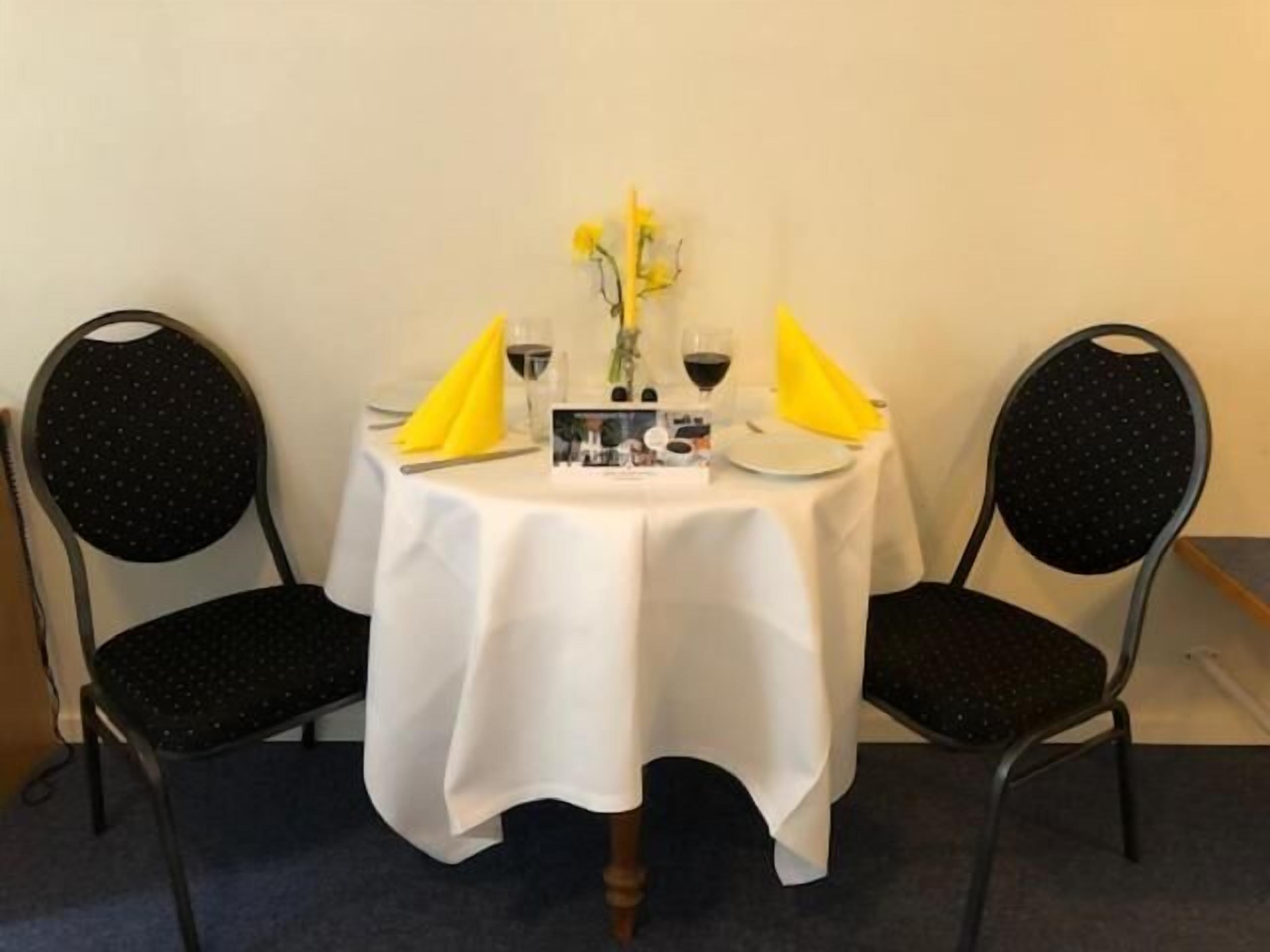 A table set for two with a white tablecloth, two chairs, a vase with yellow flowers, and a menu.