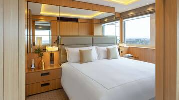 A modern hotel room with a large bed, a bedside table with a lamp, a mirror, and a view of the city.