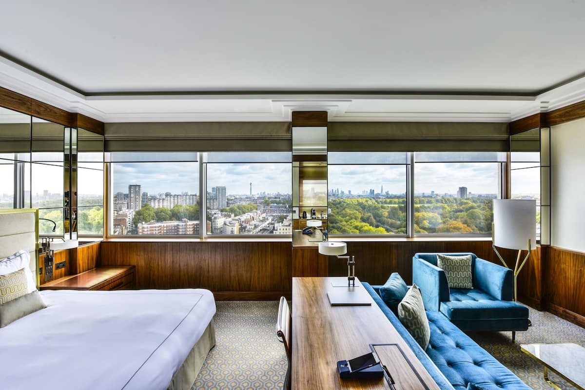 A hotel room with a large window, a bed, a sofa, a desk, and a lamp.