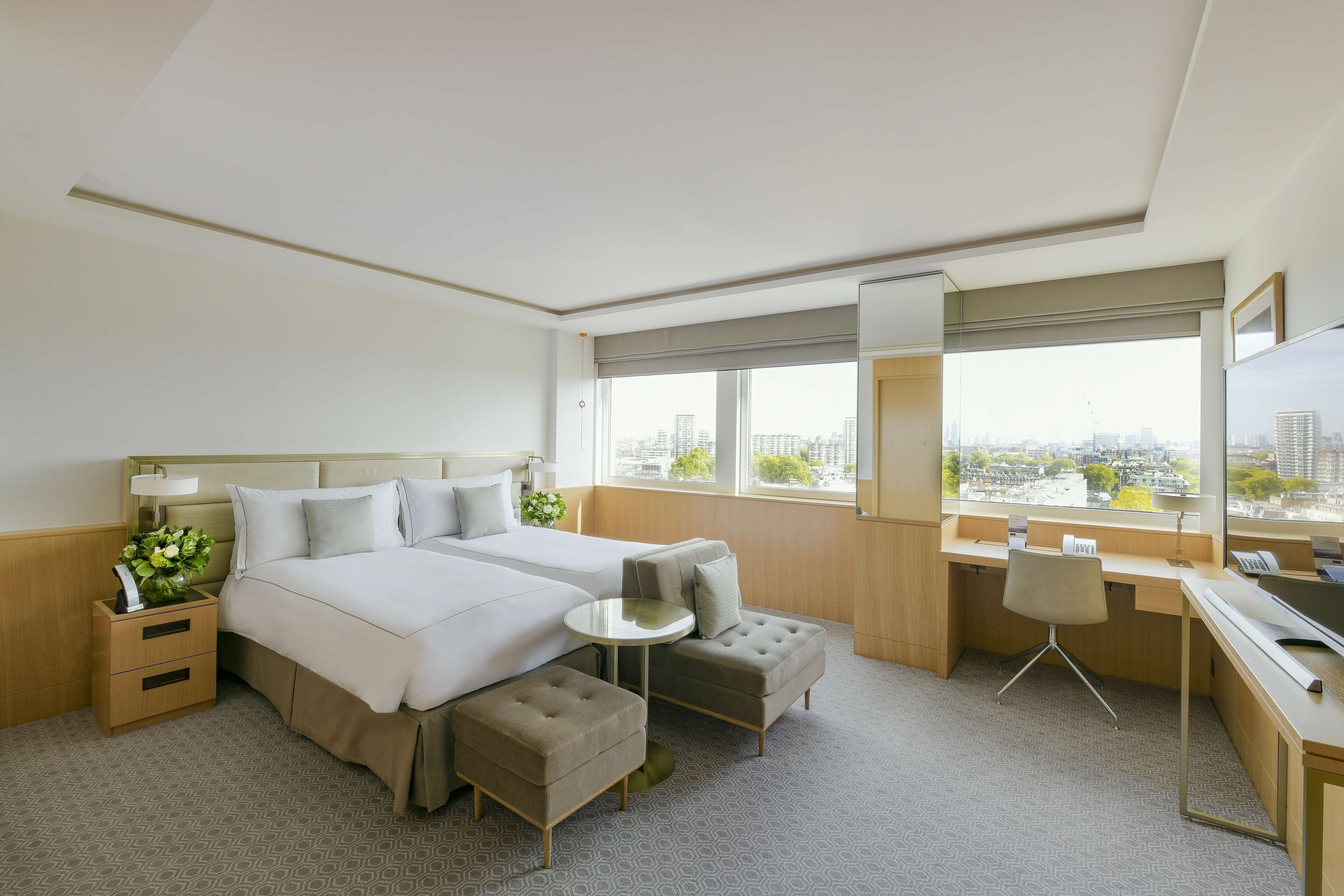 A hotel room with two beds, a desk, a chair, a small table, and a large window with a city view.