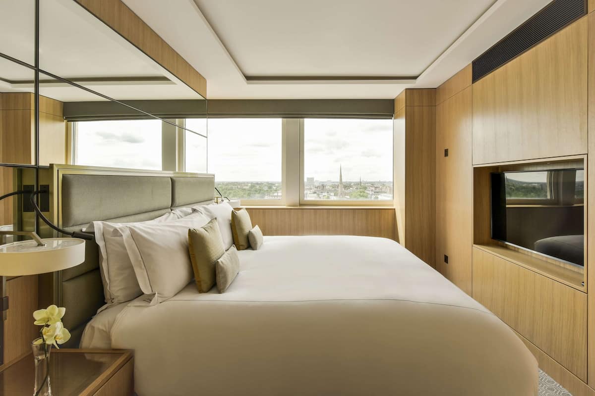 A modern hotel room with a large bed, a TV, a window with a city view, and a bedside table with a lamp and flowers.