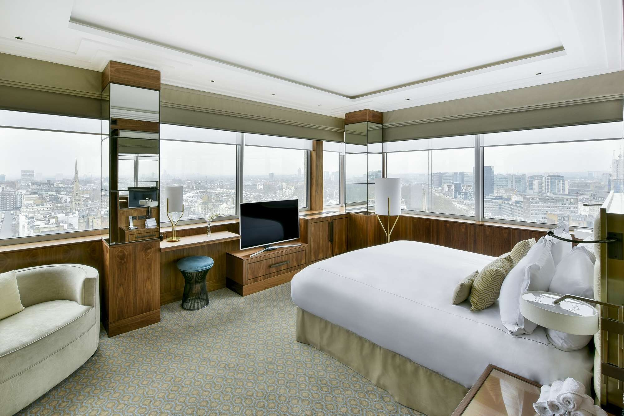A modern hotel room with a large bed, a desk with a TV, a chair, and a view of the cityscape.