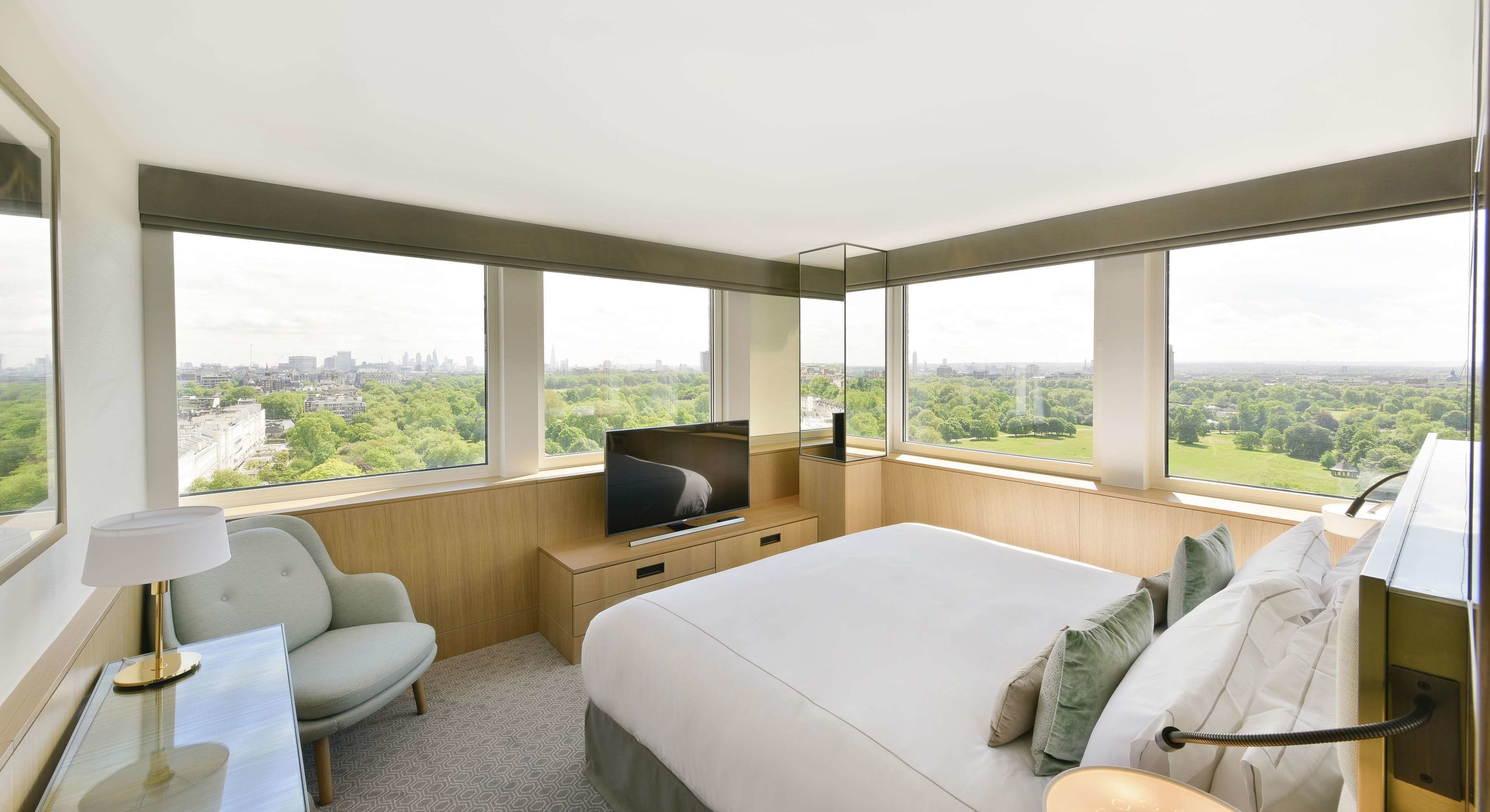 Une chambre d’hôtel moderne dotée d’un grand lit, d’une télévision, d’un fauteuil et offrant une vue sur la ville et la verdure.