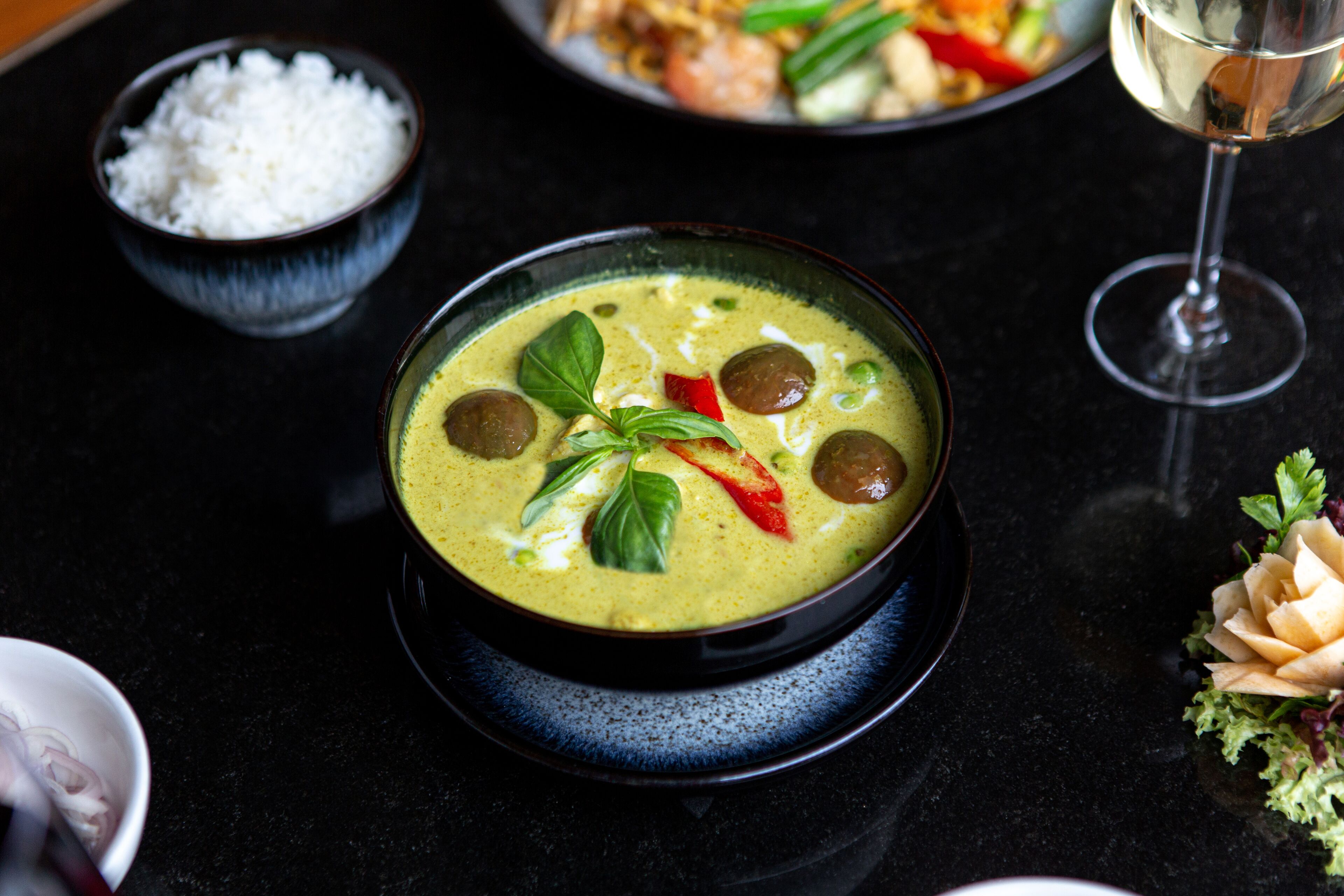 A bowl of soup with various ingredients, a glass of white wine, and a bowl of rice on a dark table.
