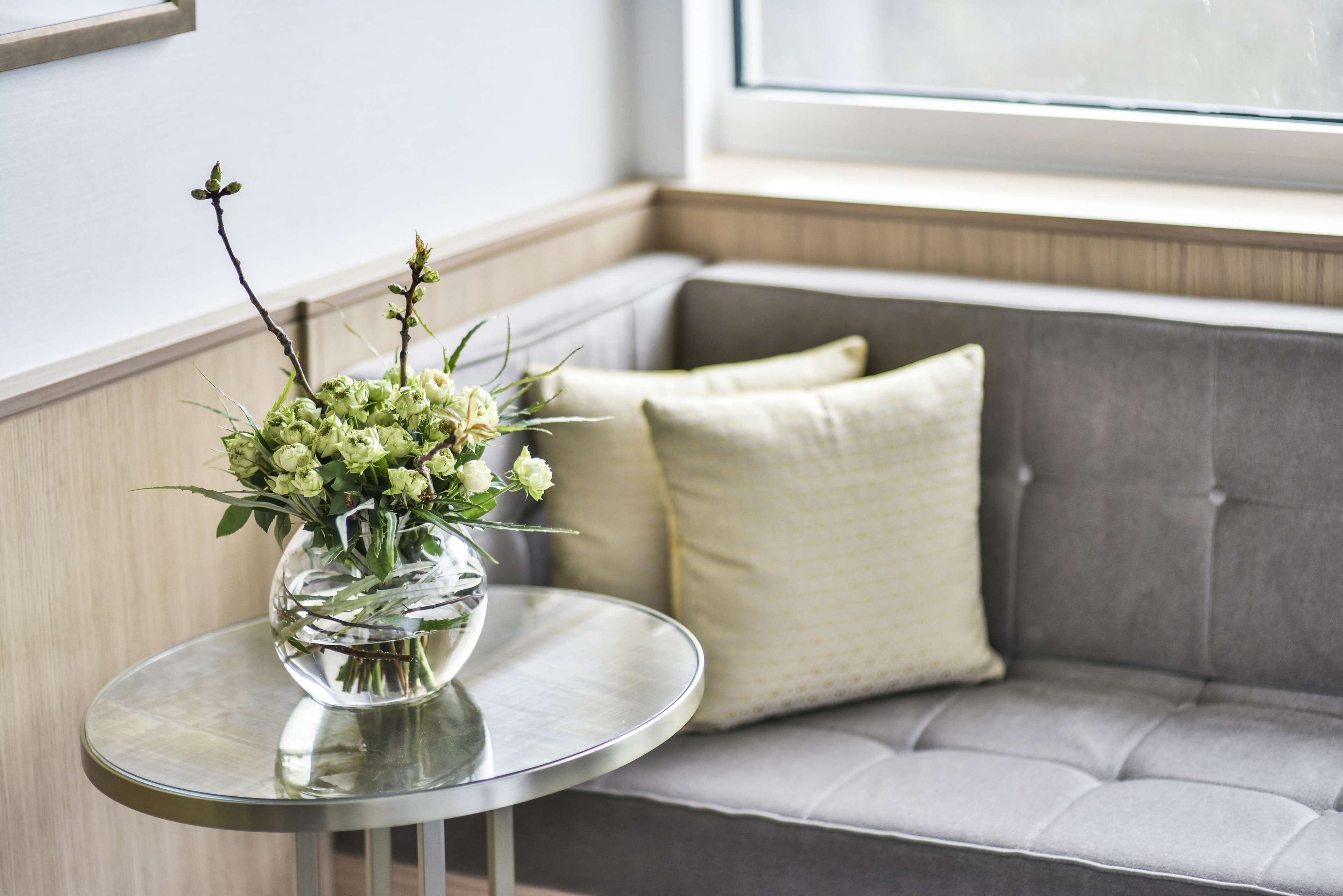 A round glass table with a vase of flowers in a clear glass bowl, placed on a grey sofa with a cushion.