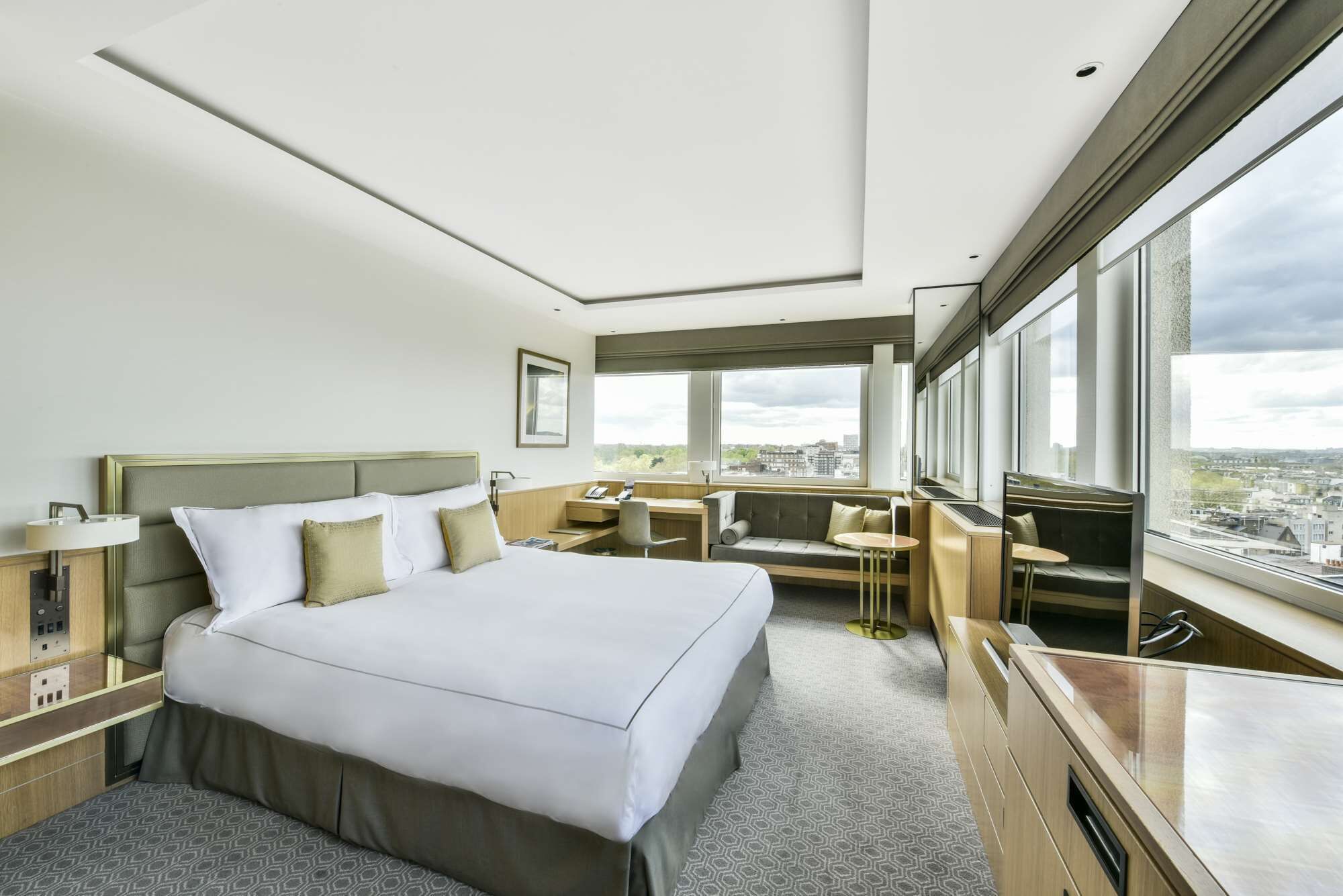 Una habitación de hotel moderna con una cama grande, un escritorio y un área de descanso junto a un ventanal con vistas a la ciudad.