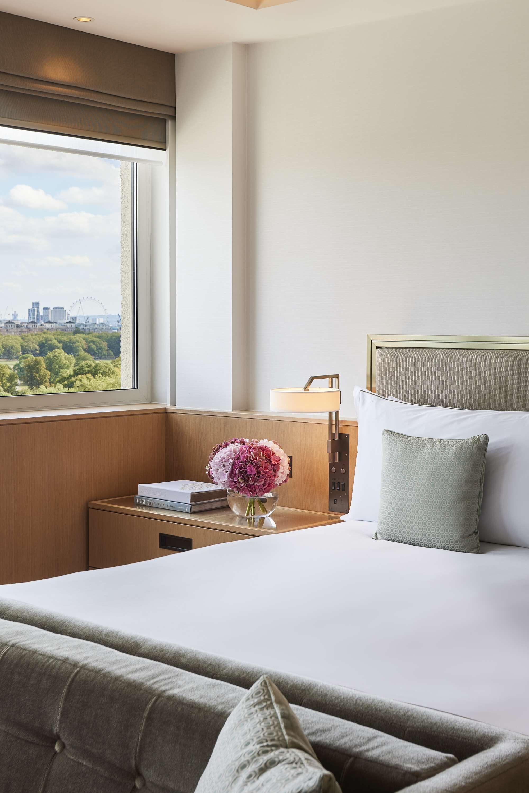 A hotel room with a large bed, a nightstand with a lamp, a vase of flowers, and a view of a cityscape through the window.