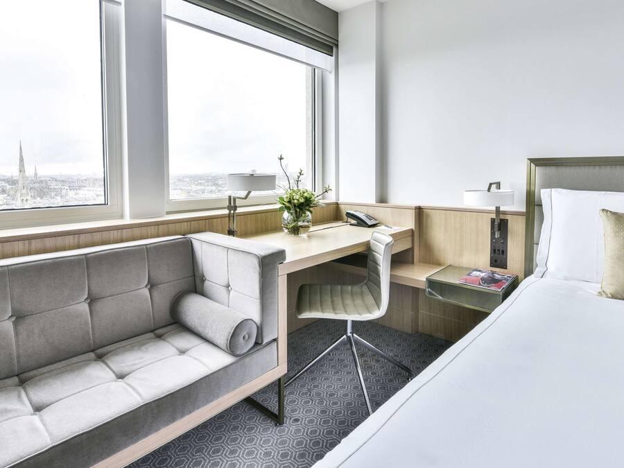 A hotel room with a bed, a desk, a sofa, and a view of a cityscape.