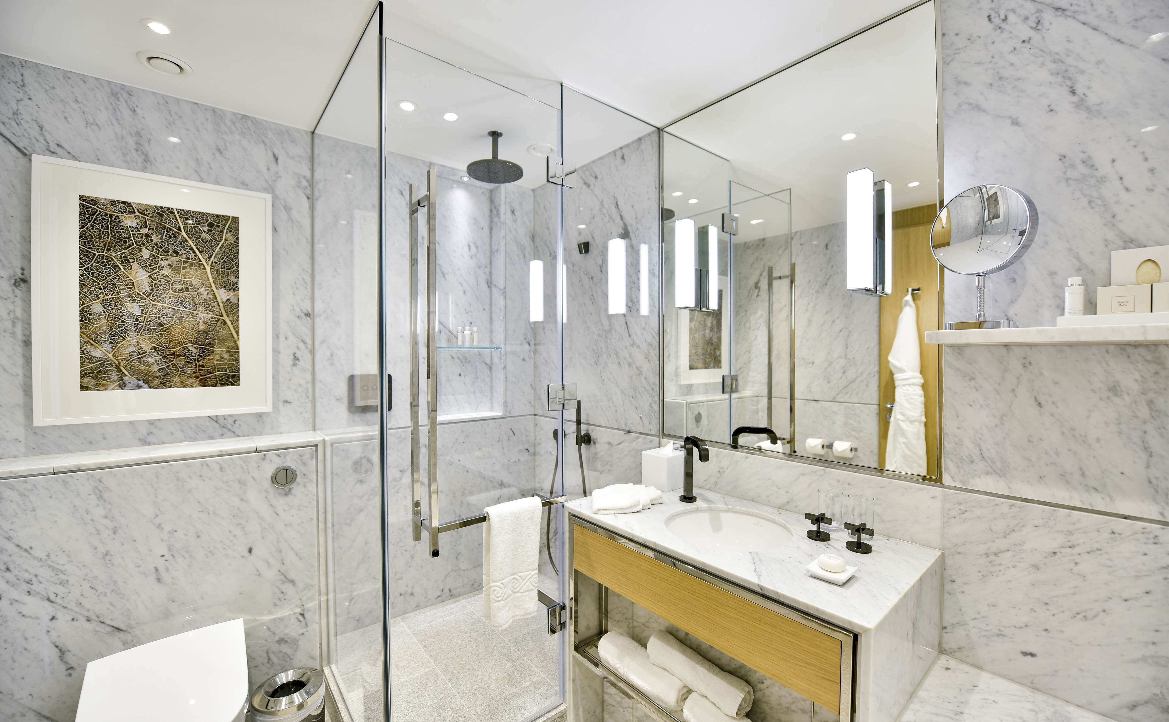 Un bagno moderno con cabina doccia in vetro, parete rivestita in marmo, uno specchio grande e un'opera d'arte incorniciata.