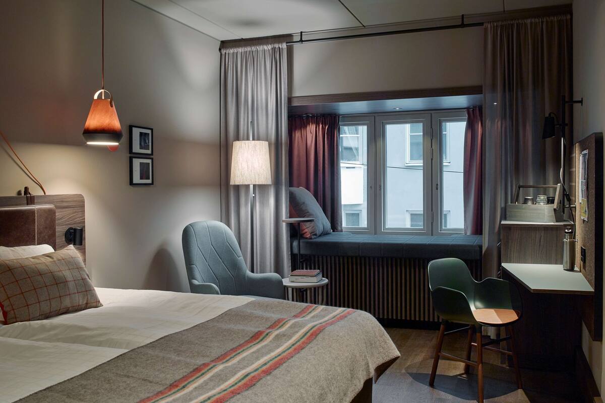 A hotel room with a bed, a desk, a chair, a window with curtains, a lamp, and a radiator.