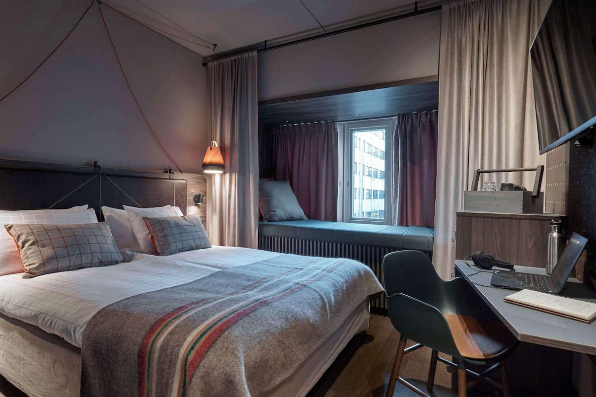 A hotel room with a bed, a desk, a chair, a window with curtains, and a radiator.
