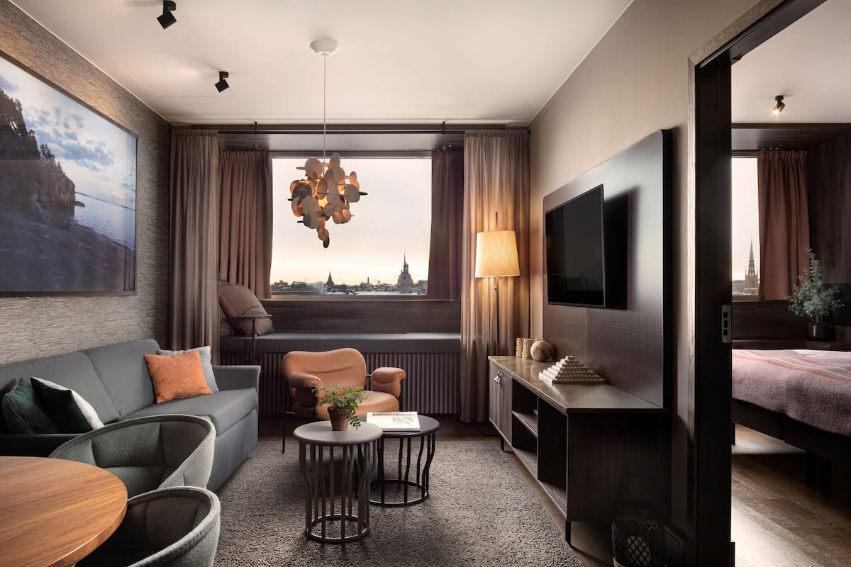 A modern hotel room with a large window, a sofa, a chair, a small round table, a TV, and a bed.