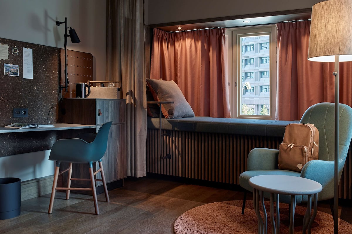 A modern hotel room with a bed, a desk, a chair, and a view of the city.