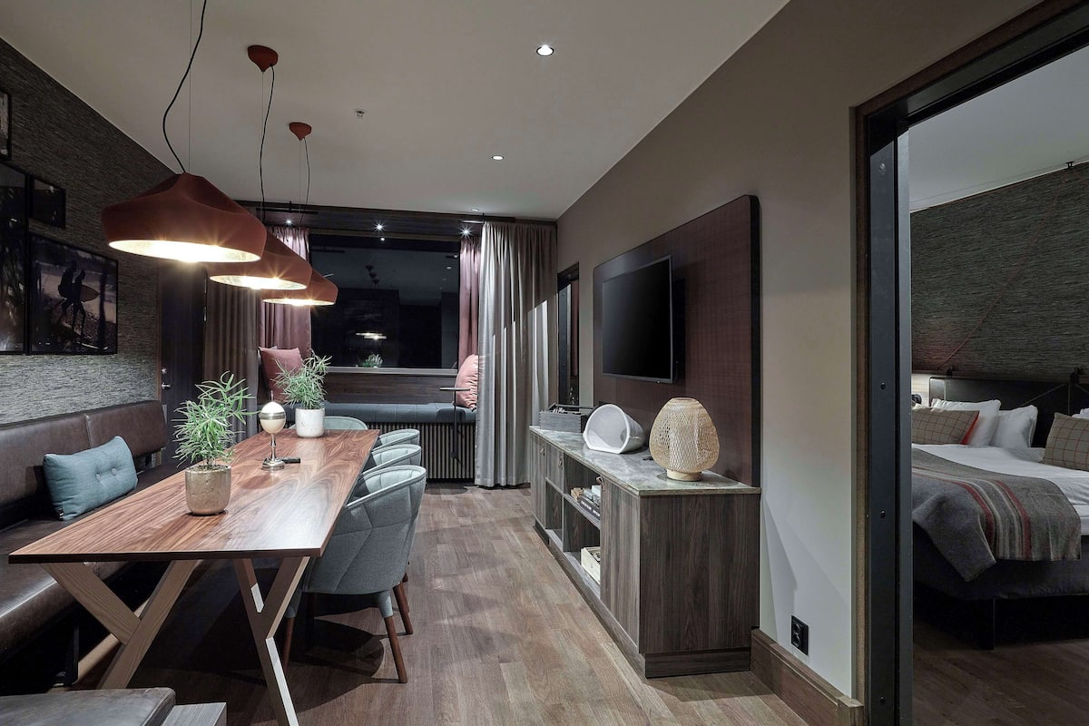 A modern living room with a wooden dining table, grey upholstered chairs, a large flat-screen TV, and a cozy seating area with a sofa and cushions.