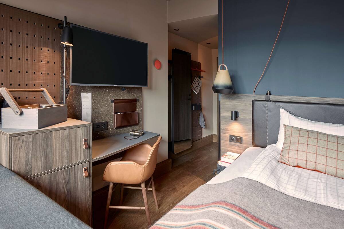 A modern hotel room with a desk, chair, bed, and a TV mounted on the wall.