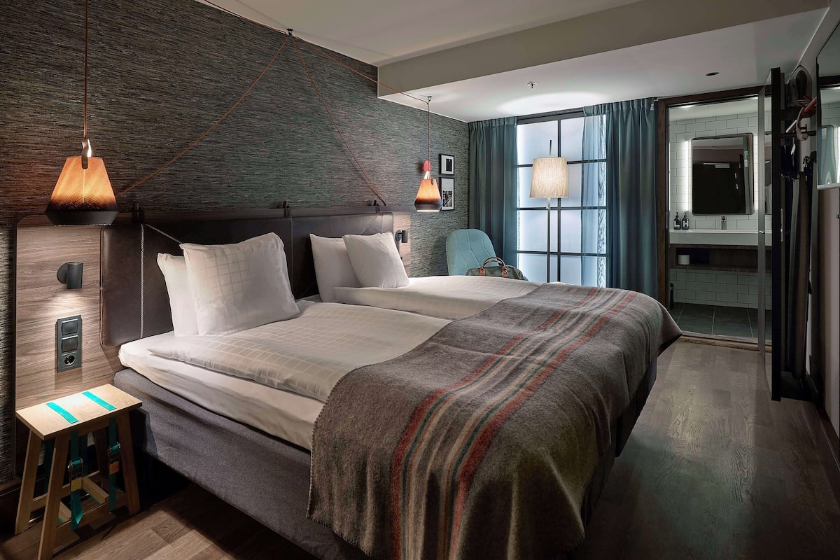 A modern hotel room with a large bed, a bedside table, a chair, a large window with curtains, and a bathroom visible through an open door.
