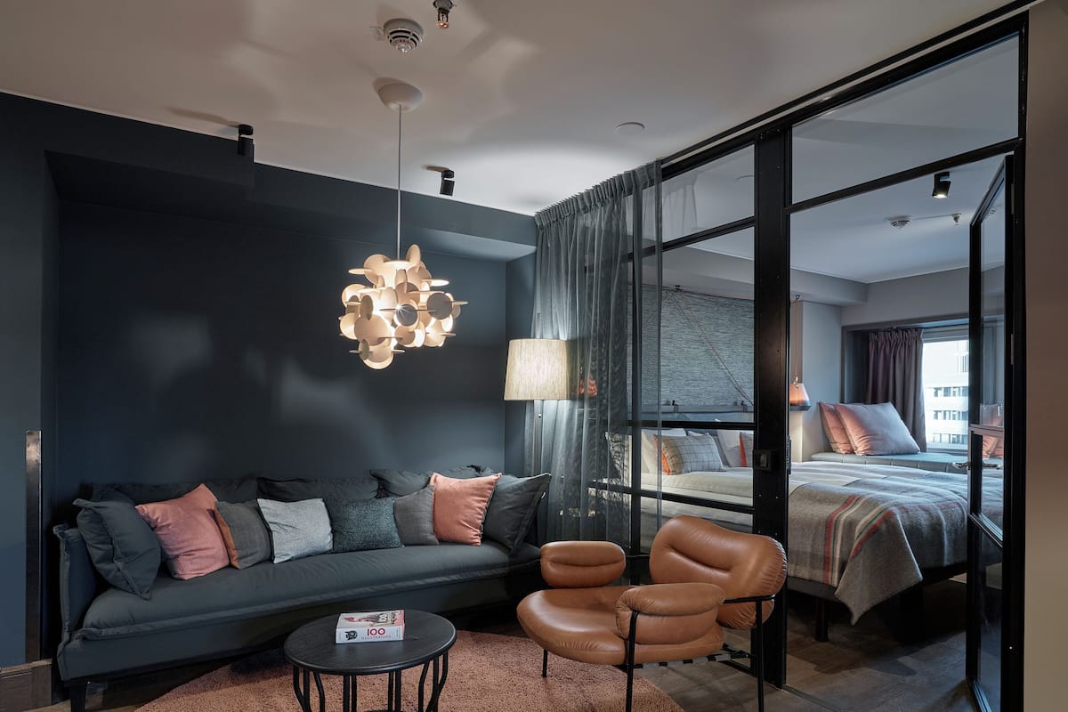 A modern hotel room with a dark blue accent wall, a sofa with multiple cushions, a leather armchair, a round coffee table, and a bed with a view of the city.
