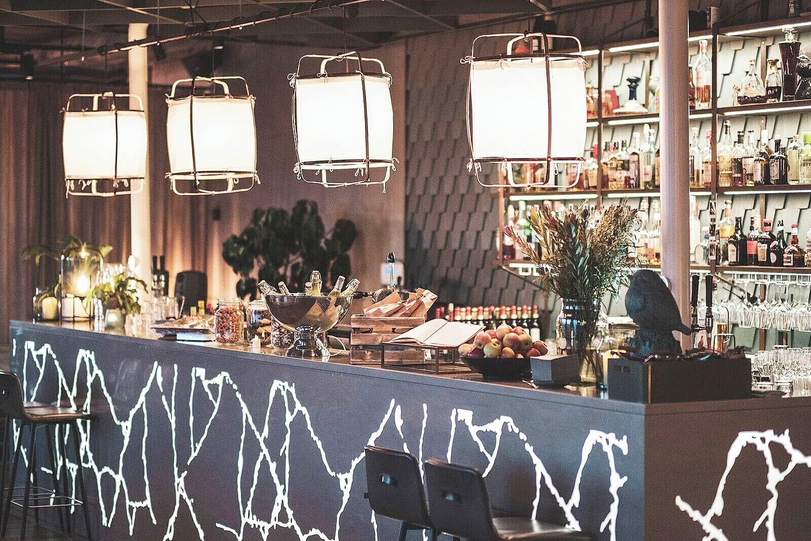 Un espace bar agrémenté de luminaires suspendus, d’un comptoir et d’étagères remplies de bouteilles et de verres.