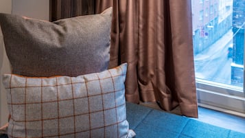 A bed with a blue mattress and two pillows, one with a checkered pattern and the other plain, next to a window with brown curtains.