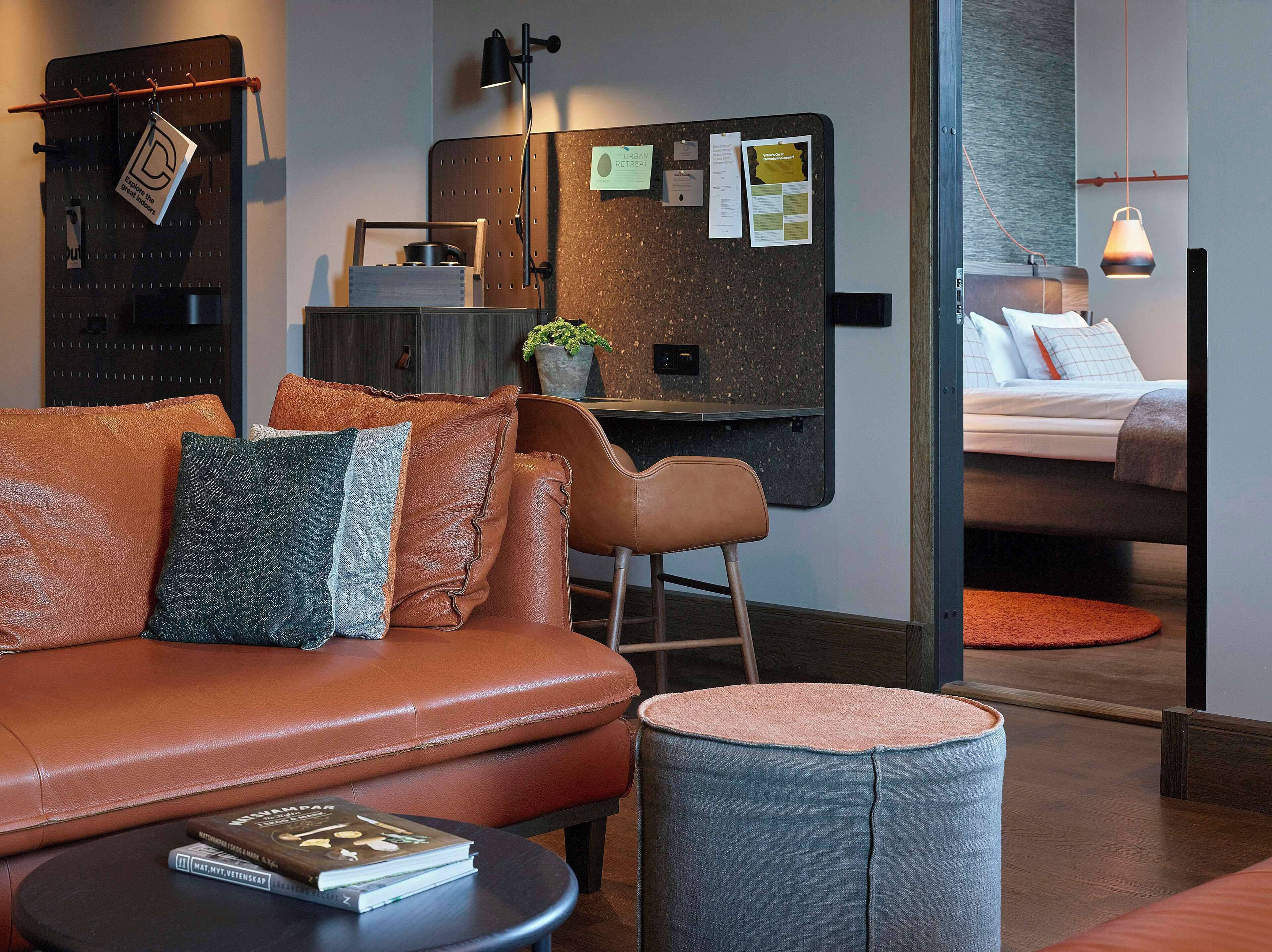 Een moderne hotelkamer met een leren bank, een klein rond poufje, een bureau met een potplant, een bed met kussens en een prikbord met berichten.