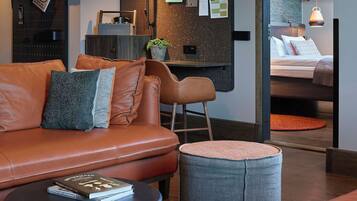 Een moderne hotelkamer met een leren bank, een klein rond poufje, een bureau met een potplant, een bed met kussens en een prikbord met berichten.