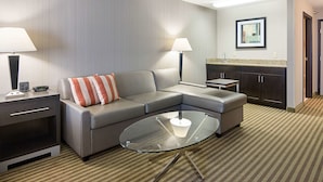 Une chambre d’hôtel moderne avec un canapé d’angle gris, une table basse en verre, une petite table d’appoint, une lampe, une œuvre d’art encadrée au mur et un coin cuisine.