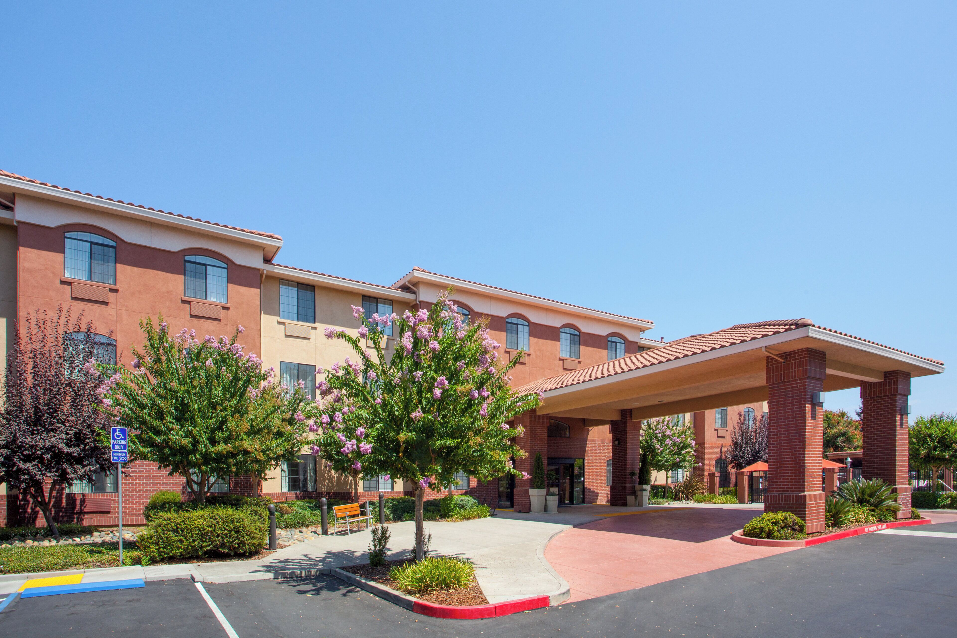 A multi-story hotel with a covered entrance, landscaped gardens, and a parking area.