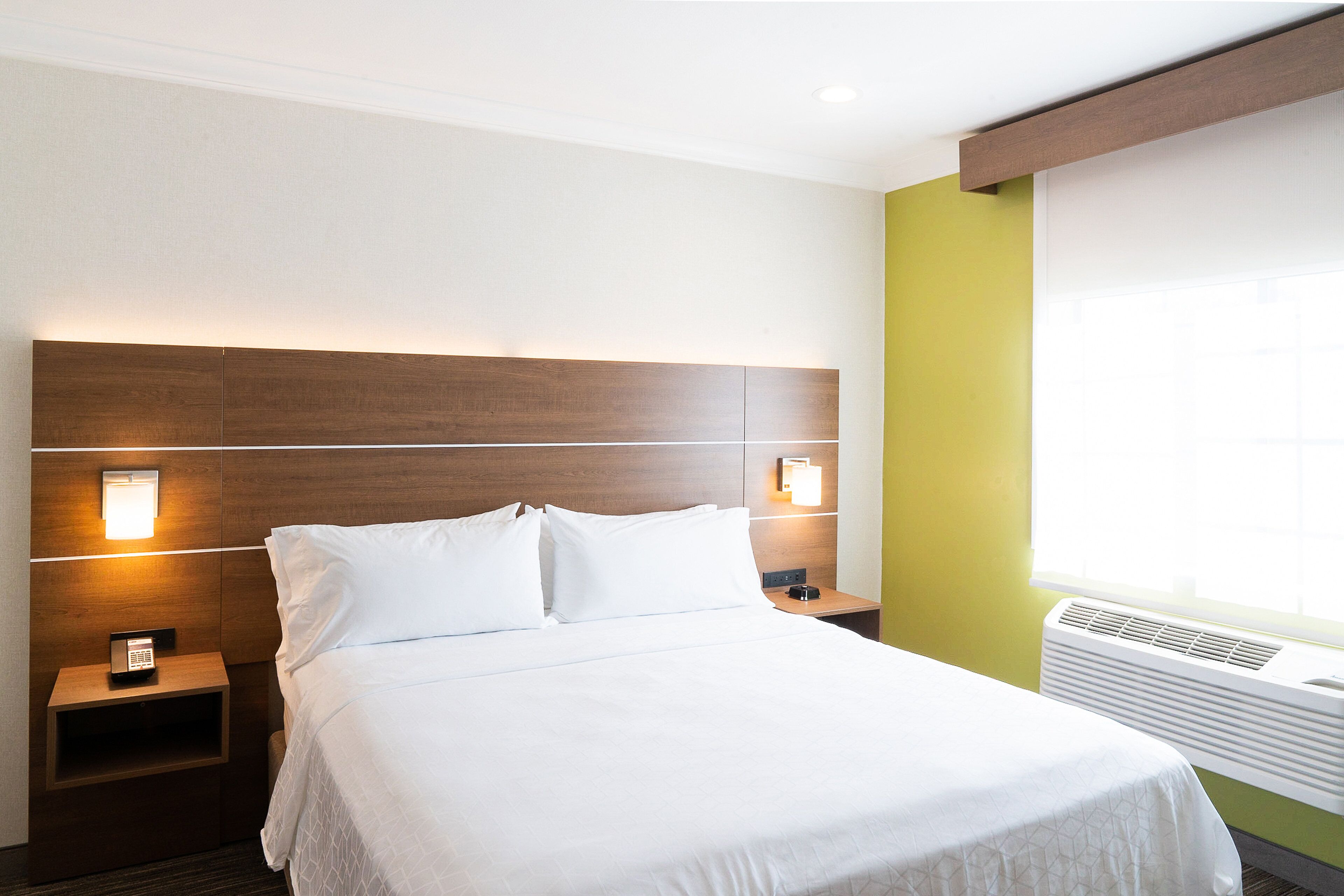 A hotel room with a bed, bedside tables, a nightstand, a wall-mounted phone, and a window with an air conditioning unit.