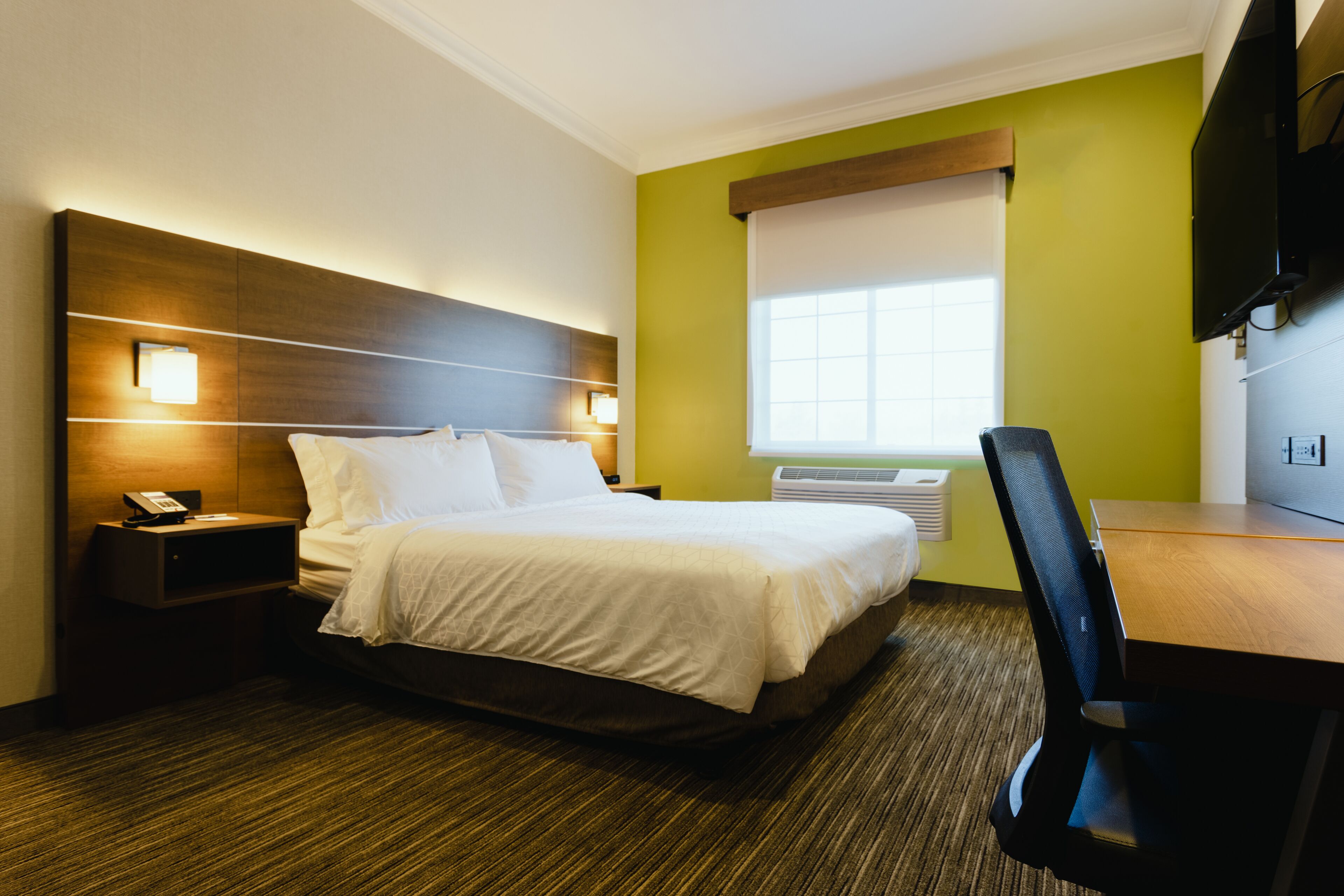 A hotel room with a bed, bedside table with a phone, a desk with a chair, a television, and a window with blinds.