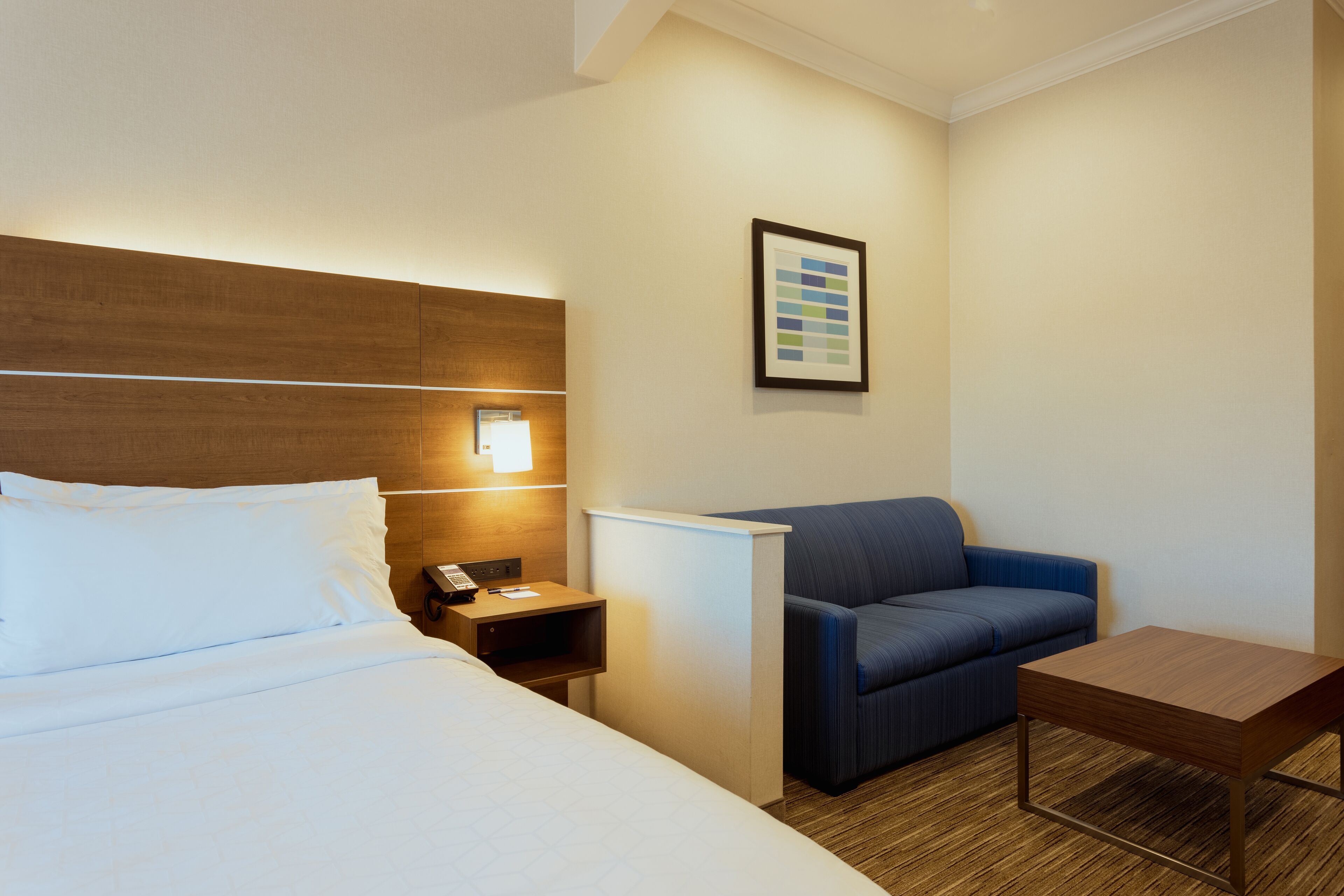 A hotel room with a bed, a nightstand with a phone, a blue armchair, and a small wooden table.