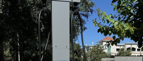 Una estación de carga para vehículos eléctricos con varios cables conectados, con un fondo de árboles y un edificio.