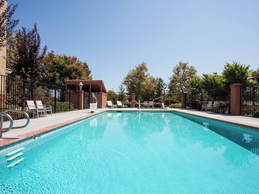A rectangular swimming pool with clear blue water, surrounded by a low brick wall and metal railings. There are lounge chairs and a covered seating area with a roof.