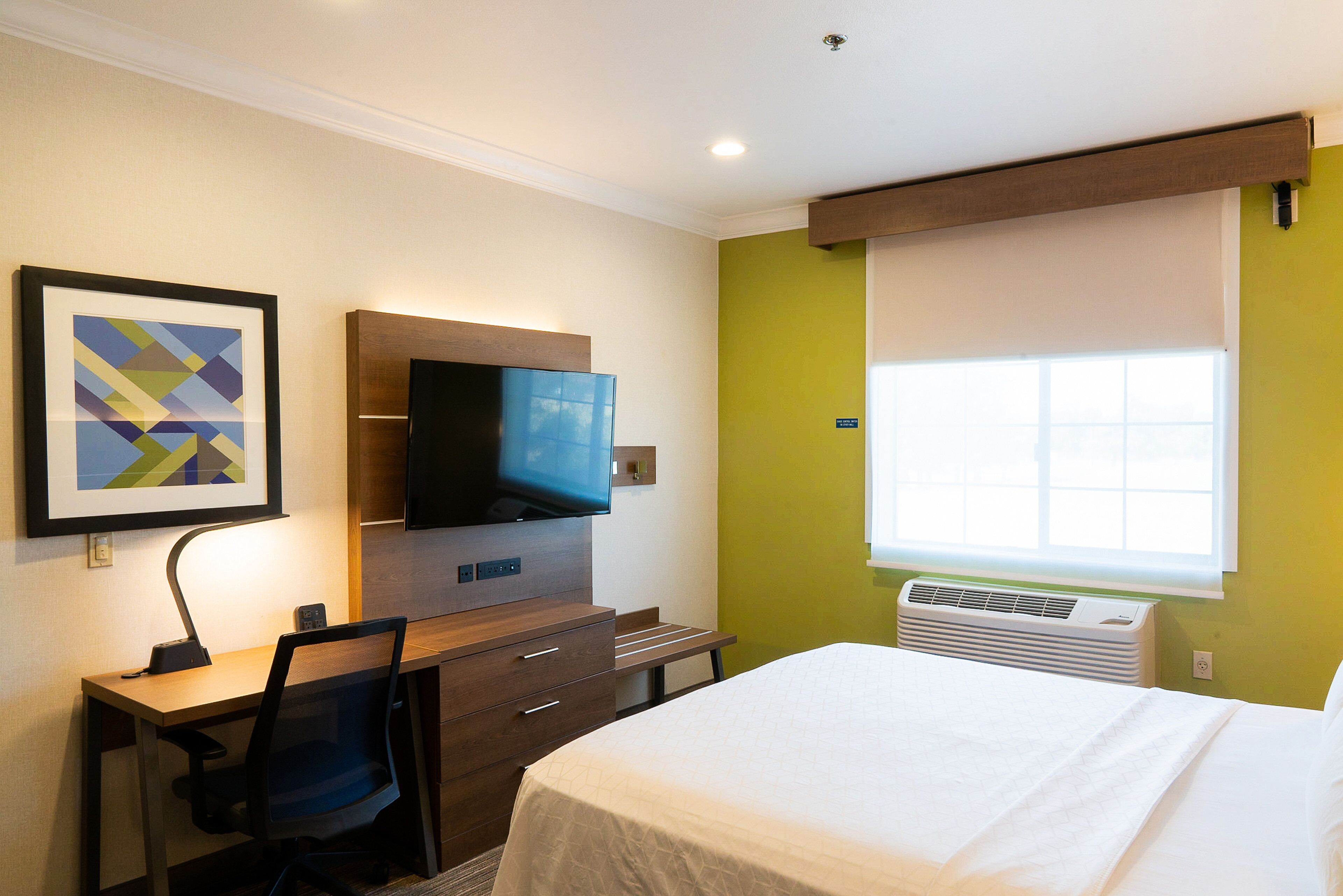 Habitación de hotel con cama, escritorio, silla, televisor y ventana con persianas.