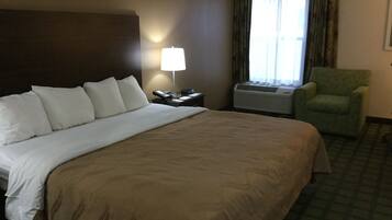 A hotel room with a large bed, a nightstand with a lamp, an air conditioning unit, and a green armchair.